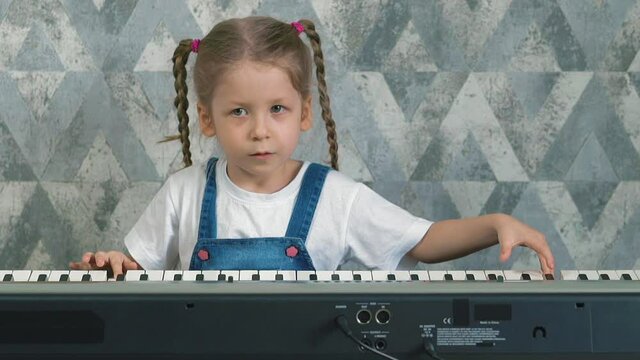 slow motion portrait of little girl at home in quarantine play on electric piano sad slow song, child in blue sundress, white T-shirt, with long blond hair braided in pigtails