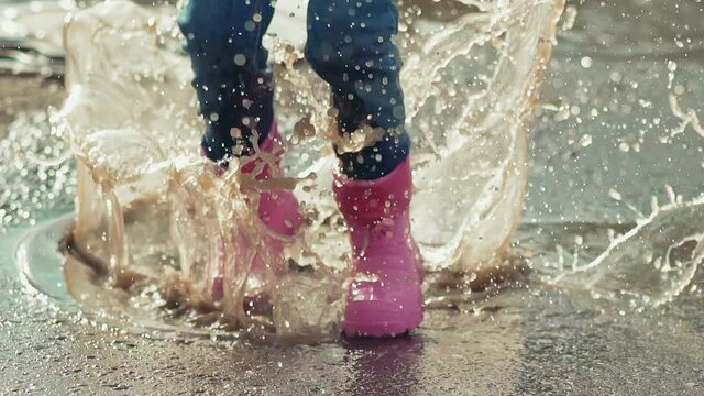 closeup slow motion little child runs and jumps into deep puddle in rainboots and jeans with all over span, picking up large splash fountain, soiling clothes, and wet pants, in sunny weather