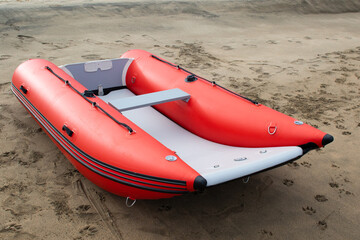 An inflatable boat on the beach it is red in color with a grey floor & black stripes. It is a...