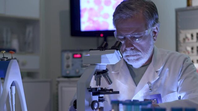A cancer research scientist in a laboratory looking through a microscope with a display of cancer cells on the monitor in the background.