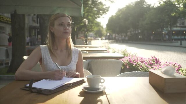 Young blonde female in white dress in outside cafe terrace table writing thoughts in personal diary journal looking away