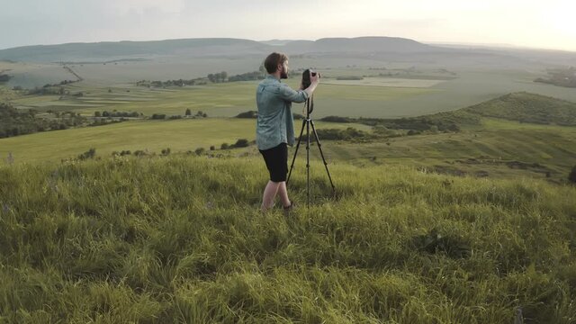 Professional photographer in summer outfit using tripod and digital camera for making pictures of amazing mountains. Mature man standing on hill with green nature around.