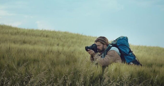 Competent photographer with big backpack standing among green field and taking pictures of spring nature. Mature man using professional digital camera for creating amazing photos outdoors.