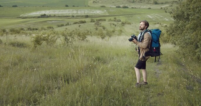 Professional photographer standing on hill and making pictures with background of beautiful green nature. Bearded man with big backpack enjoying favorite hobby on fresh air.