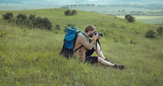Mature man in casual outfit sitting high on green hill and making photos on professional camera. Bearded man relaxing on ground while shooting beautiful nature around.