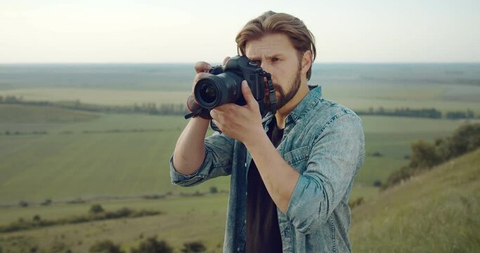 Portrait of professional bearded photographer taking pictures on digital camera while standing high on green hill. Mature man in denim shirt spending free time for favorite hobby.
