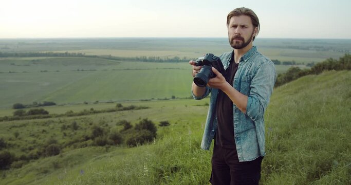 Mature bearded man in casual clothing standing on high green hill and using digital camera for making photos on fresh air. Concept of working process and photography