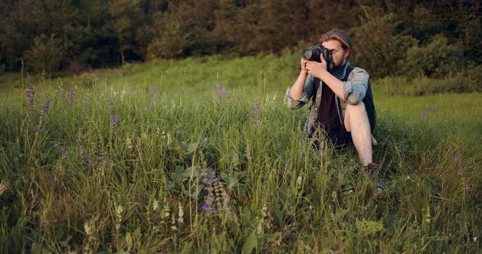 Mature bearded man with backpack sitting on green hill and using digital camera for making photos on fresh air. Concept of working process and photography