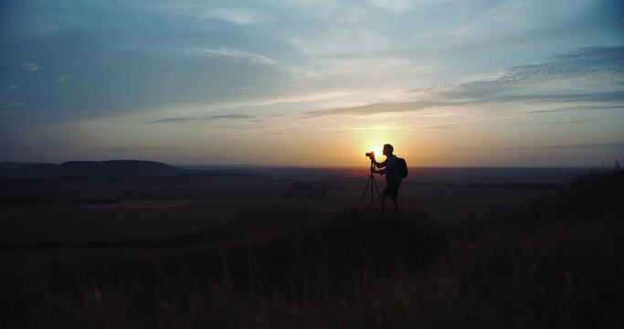 Full length portrait of professional photographer taking photos of amazing sunset and using tripod with digital camera. Dark atmosphere around.