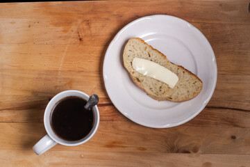 A white Cup with a spoon and coffee, on a wooden Board, a white plate with a slice of homemade bread and butter.