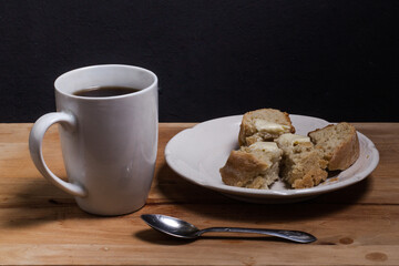 A white Cup with a spoon and coffee, on a wooden Board, a white plate with a slice of homemade bread and butter.