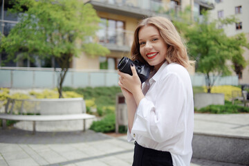 Fototapeta premium Photographer girl walks around the city with a reflex camera. A young blonde learns to take pictures. The model is looking at the camera and holding a camera in her hands. Photo Freelance Smile
