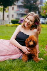 A girl in a chic long pink dress is sitting in the yard with her dog, a brown poodle. The girl walks with the dog, the hostess and the animal. A girl is playing with her dog.