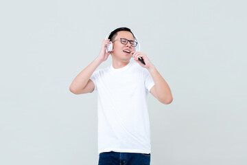 Young happy Asian man listening to music from headphones and singing isolated on light gray background