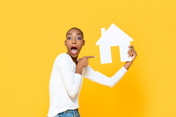 Shocked African American woman gasping and pointing to house cutout model isolated on yellow...