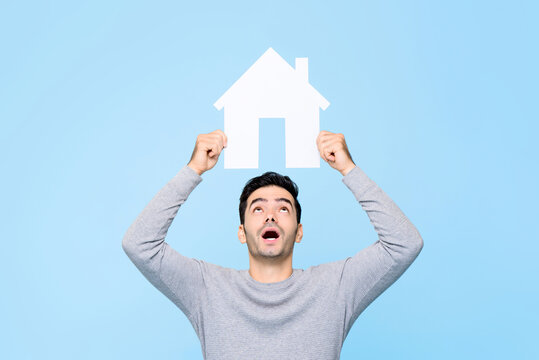 Young Man Holding House Cutout Model Overhead And Looking Up Isolated On Light Blue Background