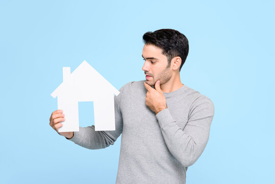 Young man looking at house model and thinking isolated on light blue background