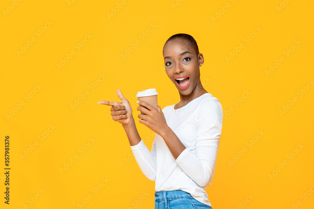 Wall mural Surprised African American woman with takeaway coffee cup smiling and pointing hand to empty space aside isolated on yellow background