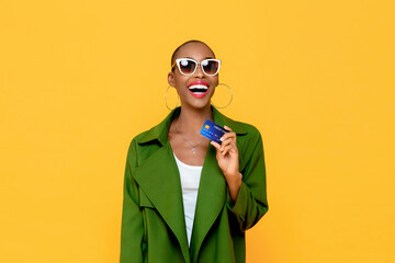 Happy fashionable African American woman smiling and showing credit card in hand on yellow isolated...