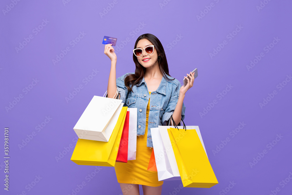 Wall mural Happy young Asian girl carrying colorful shopping bags with credit card and mobile phone in hands isolated in purple background