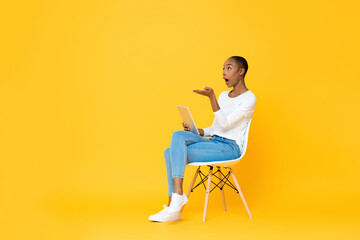 Young surprised African American woman sitting and opening hand looking up to copy space while...