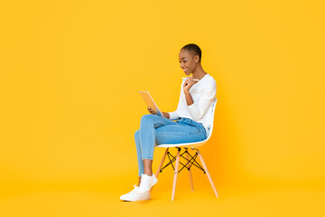 Young African American woman sitting and using tablet computer with stylus pen on isolated yellow...