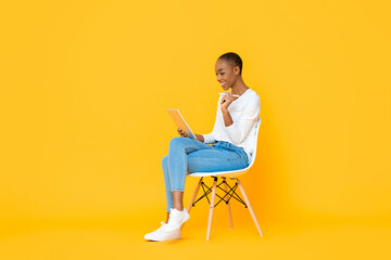 Young African American woman sitting and using tablet computer with stylus pen on isolated yellow background