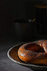 Ring shaped roll | Bagel with poppies on the top served in the dark plate. Dark teacup in the bokeh. Fresh bagel serving. Close up. Horizontal background. Copy space for the text editing. 