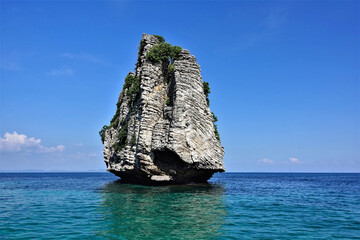 Lonely island in the Andaman Sea. A rock in the form of a pyramid rises high above the turquoise water. There are few plants on top. Reflection in water. The background is a clear bright blue sky. 