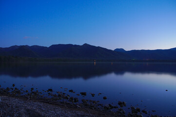 Fototapeta na wymiar 黄昏時の青い湖の風景。水面に映る空の色、遠くの山々のシルエット。