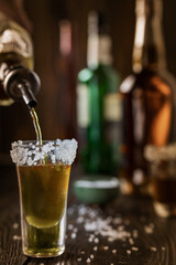 An alcoholic drink at the bar counter is poured into a tequila glass with salt at the edges, in the background are different bottles of alcohol, shallow depth of field, selective focus. The concept of