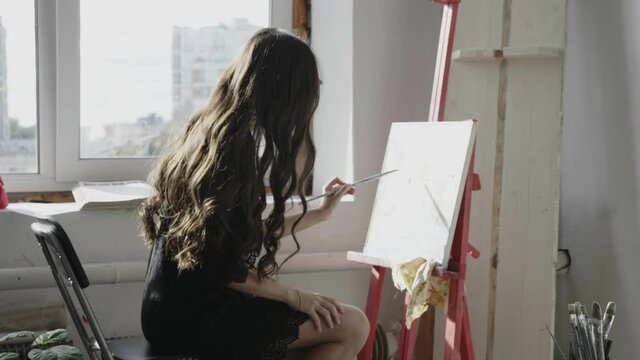 long curly haired girl in black dress draws picture with brush on easel near window in art studio at morning light slow motion