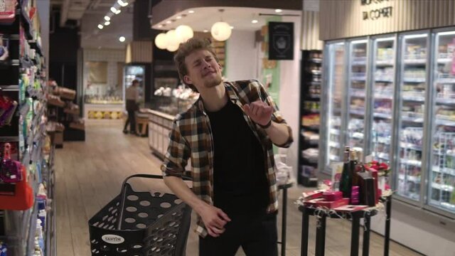 Excited young man in plaid shirt in supermarket dancing, funky moves close to shopping trolley. Positive dances in an empty food store row. Positive emotions