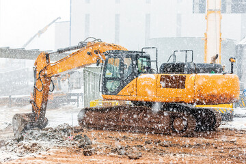 The yellow excavator is working in the construction site in snowy weather.