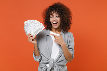 Shocked young african american woman in gray casual clothes isolated on orange background. People lifestyle concept. Mock up copy space. Pointing index finger on fan of cash money in dollar banknotes.