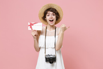 Excited tourist woman in dress hat with photo camera isolated on pink background. Female traveling to travel weekends getaway. Air flight journey concept. Hold gift certificate pointing thumb aside.