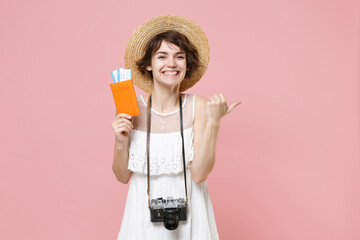 Smiling young tourist girl in summer dress hat with photo camera isolated on pink background. Traveling abroad weekend getaway. Air flight journey concept. Hold passport tickets pointing thumb aside.