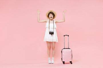 Shocked tourist girl in summer dress hat with photo camera suitcase isolated on pink wall background. Traveling abroad weekend getaway. Air flight journey concept. Keeping mouth open, spreading hands.