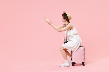 Fototapeta na wymiar Side view of tourist girl in dress hat with photo camera isolated on pink background. Female traveling abroad to travel weekend getaway. Air flight journey concept. Sit on suitcase spreading hands.