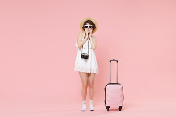 Pretty tourist girl in summer dress hat sunglasses with suitcase photo camera isolated on pink background. Traveling abroad to travel weekends getaway. Air flight journey concept. Put hands on cheeks.