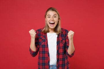 Joyful young blonde woman girl in casual checkered shirt posing isolated on red wall background studio portrait. People sincere emotions lifestyle concept. Mock up copy space. Doing winner gesture.