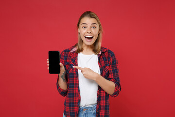 Excited young blonde woman girl in casual checkered shirt posing isolated on red background. People lifestyle concept. Mock up copy space. Point index finger on mobile phone with blank empty screen.