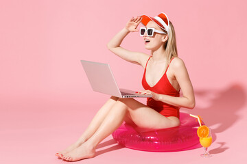 Young woman in red one-piece swimsuit cap sunglasses isolated on pink background. People summer vacation rest concept. Sit in swim inflatable ring work on laptop pc computer looking far away distance.