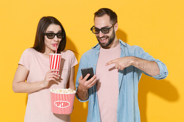 Funny young couple friends guy girl in 3d glasses isolated on yellow background. People in cinema concept. Watching movie film, hold bucket of popcorn, cup of soda point index finger on mobile phone.