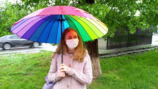 Caucasian girl in a protective mask walks under an umbrella on an empty street in spring rain. Safety and social distance during a coronavirus pandemic. New normal, the implications of quarantine