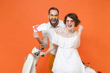 Happy young couple friends guy girl in white summer clothes glasses sit on moped isolated on orange background. Driving motorbike transportation concept. Hold gift certificate doing winner gesture.