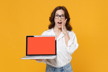 Shocked young business woman in white shirt glasses isolated on yellow background studio. Achievement career wealth business concept. Hold laptop pc computer with blank empty screen put hand on cheek.