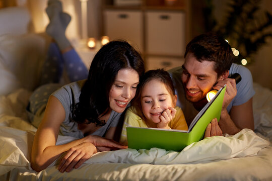 People And Family Concept - Happy Mother, Father And Little Daughter Reading Book With Torch Light In Bed At Night At Home