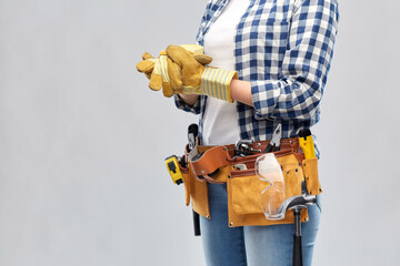 Fototapeta na wymiar repair, construction and building concept - woman or builder with helmet and working tools on belt putting protective gloves on over grey background