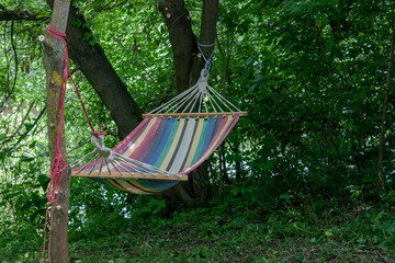 A multi-colored hammock hangs between trees in the shade. Rest on a fabric hammock on a day off. Rainbow hammock among the green trees.